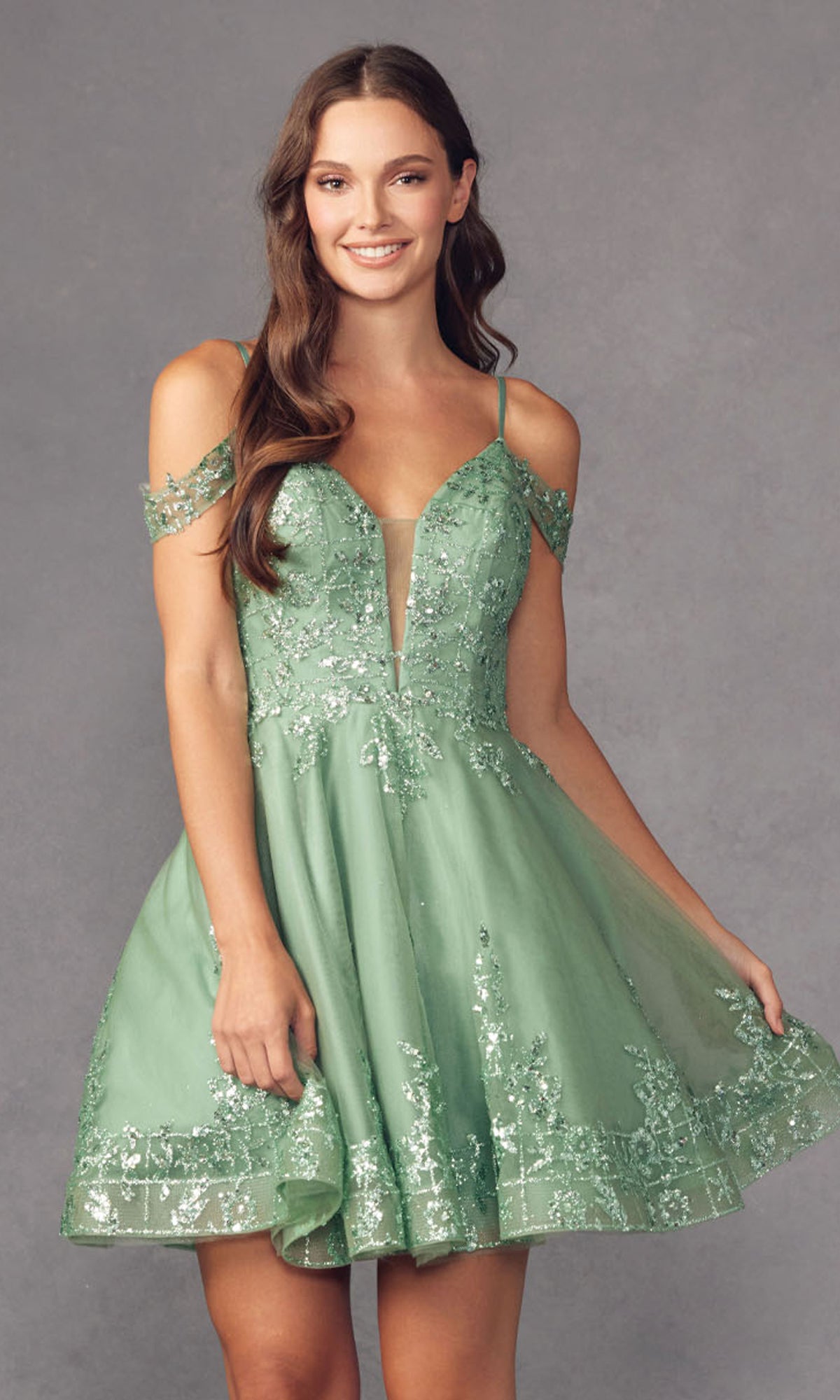 A woman smiles and holds the edge of her dress while wearing the Glitter-Print Short Babydoll Homecoming Dress 903, a short light green homecoming style with sequin embellishments, standing against a gray background.